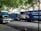 Several moving trucks from Divine Moving & Storage Ltd are parked along a street lined with trees. The trucks are large and blue, with company logos and contact information visible. A painting detail is incorporated into the truck design. The street has a sidewalk with some water reflecting the surroundings.