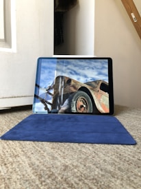 A tablet displaying an image of a rusty, old vehicle with a clear blue sky in the background. The tablet is placed on a blue case, which rests on a carpeted floor. A white door and part of a wooden staircase are visible in the surrounding environment.
