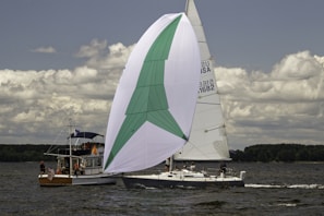 Two sailboats are gliding on a large body of water. The larger boat features a prominent white and green sail, while the smaller vessel is equipped with a motor and carries several passengers. The clouds above are scattered, and the horizon is lined with trees.
