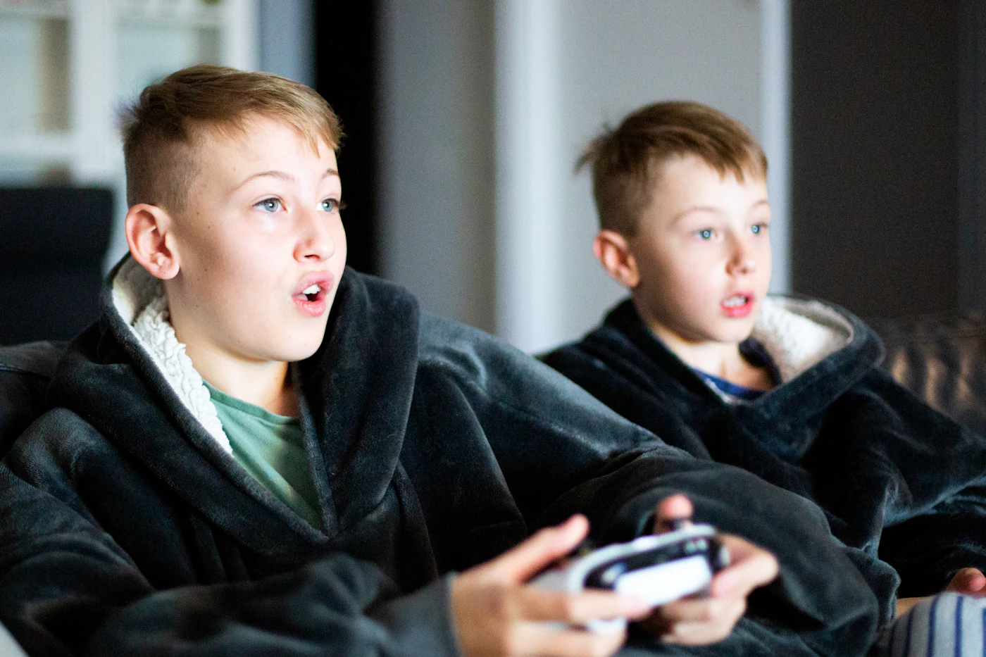Two boys sitting side by side on a couch, both holding game controllers.