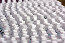 Rows of identical plastic water bottles are closely arranged, creating a pattern of white caps against clear bottles. The bottles are tightly packed, emphasizing uniformity and mass production.