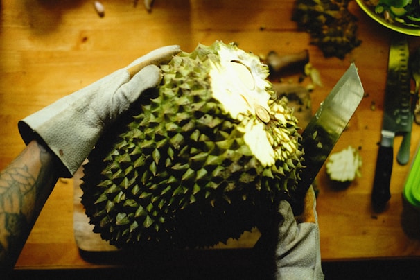 A person wearing protective gloves is preparing a durian fruit on a wooden table. The spiky durian is being cut open with a large knife. There are other kitchen tools and a partially cut durian in the background.