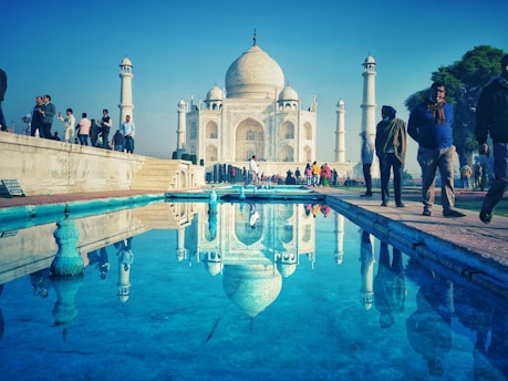 The iconic Taj Mahal is reflected in a clear pool of water with a vivid blue hue. Numerous visitors are walking along the pathway or standing nearby, admiring the monument. The sky is clear, enhancing the grandeur of the white marble structure and its minarets, which stand tall against the sky.