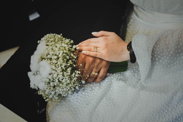 A close-up of a bride and groom's hands showing wedding rings. The bride holds a bouquet of small white flowers while wearing a white textured dress. The groom's hand is underneath, with a black suit visible in the background.