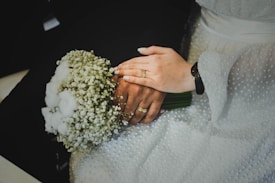 A close-up of a bride and groom's hands showing wedding rings. The bride holds a bouquet of small white flowers while wearing a white textured dress. The groom's hand is underneath, with a black suit visible in the background.