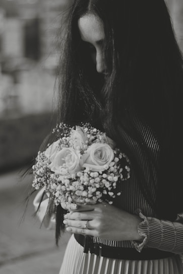 A black and white photograph of a person with long dark hair holding a bouquet of flowers, primarily roses and baby's breath, against a soft focus background. The person is looking down, creating a contemplative atmosphere.
