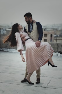 A man and woman are engaged in a dance pose on a rooftop with an urban background. The woman is being dipped by the man, wearing an elegant pleated pink skirt and black heels. The man is dressed in a formal outfit with a beige vest and dark blue shirt.
