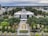 A grand architectural structure with a large dome is set amidst a lush green landscape. It is surrounded by tall trees, palm trees, and well-maintained gardens. The building's classical architectural style features columns and intricate details. The surroundings include roadways and modern city buildings, suggesting an urban environment.