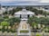 A grand architectural structure with a large dome is set amidst a lush green landscape. It is surrounded by tall trees, palm trees, and well-maintained gardens. The building's classical architectural style features columns and intricate details. The surroundings include roadways and modern city buildings, suggesting an urban environment.