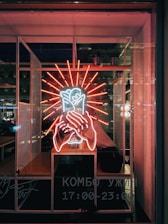 A neon sign featuring a vibrant red and white illuminated design, depicting hands holding a heart, is displayed in a dimly lit window. Inside, a person is seated behind the glass, partially visible surrounded by the glow of the neon. The background includes urban elements and some potted plants, with text in Cyrillic indicating business hours.