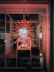 A neon sign featuring a vibrant red and white illuminated design, depicting hands holding a heart, is displayed in a dimly lit window. Inside, a person is seated behind the glass, partially visible surrounded by the glow of the neon. The background includes urban elements and some potted plants, with text in Cyrillic indicating business hours.