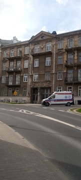 A multi-story building with an ornate facade featuring multiple balconies. There is a medical emergency vehicle parked on the road in front of the building. The road curves around the building, with some greenery visible at the edge of the road. The sky is partly cloudy.