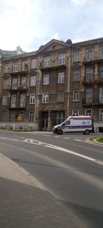 A multi-story building with an ornate facade featuring multiple balconies. There is a medical emergency vehicle parked on the road in front of the building. The road curves around the building, with some greenery visible at the edge of the road. The sky is partly cloudy.