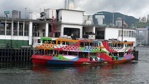 A colorful ferry with vibrant patterns docks in a harbor with modern skyscrapers in the background. The boat is adorned with life rings and has the text 'Asia's World City' on its side. A pier with a pedestrian and various urban buildings is visible.
