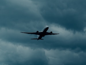 An airplane flies high in a cloudy sky, surrounded by dark, moody clouds. The silhouette of the aircraft is distinctly visible against the backdrop of thick, stormy clouds, suggesting turbulent weather conditions.
