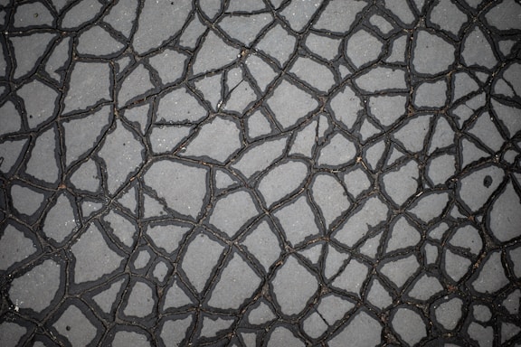 A close-up view of a cracked surface, possibly a pavement or asphalt, with irregular patterns formed by the cracks. The surface is primarily gray, and the cracks are dark, giving a distinct web-like appearance.