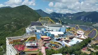 An amusement park set against a backdrop of lush green mountains and partially cloudy skies. There are various rides and attractions, including a roller coaster with bright yellow tracks and a large structure resembling a clown face. The area is bustling with visitors, and there are colorful tents, umbrellas, and buildings scattered throughout.