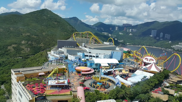 An amusement park set against a backdrop of lush green mountains and partially cloudy skies. There are various rides and attractions, including a roller coaster with bright yellow tracks and a large structure resembling a clown face. The area is bustling with visitors, and there are colorful tents, umbrellas, and buildings scattered throughout.