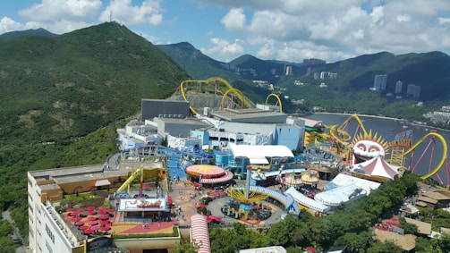An amusement park set against a backdrop of lush green mountains and partially cloudy skies. There are various rides and attractions, including a roller coaster with bright yellow tracks and a large structure resembling a clown face. The area is bustling with visitors, and there are colorful tents, umbrellas, and buildings scattered throughout.