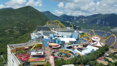 An amusement park set against a backdrop of lush green mountains and partially cloudy skies. There are various rides and attractions, including a roller coaster with bright yellow tracks and a large structure resembling a clown face. The area is bustling with visitors, and there are colorful tents, umbrellas, and buildings scattered throughout.