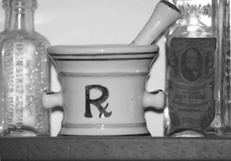 A black-and-white image featuring a mortar and pestle with the pharmacy symbol 'Rx' on its side. Flanking the mortar are two vintage glass bottles with embossed lettering, one with a label showcasing an old-fashioned portrait.
