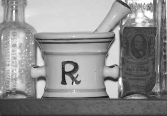 A black-and-white image featuring a mortar and pestle with the pharmacy symbol 'Rx' on its side. Flanking the mortar are two vintage glass bottles with embossed lettering, one with a label showcasing an old-fashioned portrait.