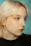 A person with light blonde hair and a nose ring is shown with multiple rhinestones or gems applied to their face. The background is a soft teal color, and the person is wearing a black top. The makeup is subtle with a focus on the eyes, enhanced by winged eyeliner.