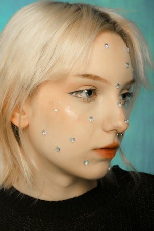 A person with light blonde hair and a nose ring is shown with multiple rhinestones or gems applied to their face. The background is a soft teal color, and the person is wearing a black top. The makeup is subtle with a focus on the eyes, enhanced by winged eyeliner.