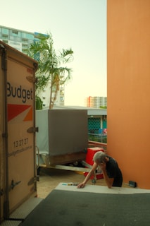 A person is leaning against a table beside a moving truck with visible branding, appearing to be engaged in a phone call. The setting includes a tropical tree, high-rise buildings in the background, and an orange wall to the right. Another parked trailer is in view, and various items are scattered on the surface in front of the individual.