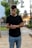 A young man is standing outdoors, looking down at his watch on his wrist. He is wearing a black t-shirt and blue jeans. The background features blurred greenery and architectural elements, suggesting a park or urban setting.