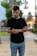 A young man is standing outdoors, looking down at his watch on his wrist. He is wearing a black t-shirt and blue jeans. The background features blurred greenery and architectural elements, suggesting a park or urban setting.