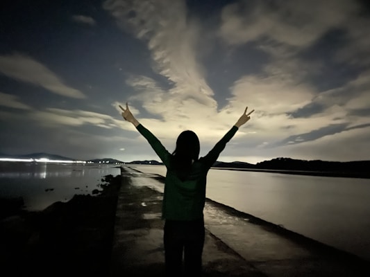 A person stands on a pathway by a body of water with arms raised in a peace sign against a backdrop of a cloudy night sky. Their silhouette is framed by the dramatic lighting of the clouds and reflections on the water. The scene is serene with distant lights visible along the horizon.