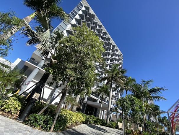A modern building with a unique geometric facade is surrounded by lush greenery and palm trees under a bright blue sky. The architecture is characterized by a series of balconies with angular designs.