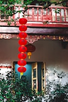 Red lanterns with Chinese characters hang vertically, surrounded by lush green plants and traditional architectural elements. A wooden window with intricate designs is partially open.