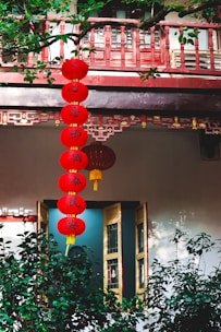 Red lanterns with Chinese characters hang vertically, surrounded by lush green plants and traditional architectural elements. A wooden window with intricate designs is partially open.