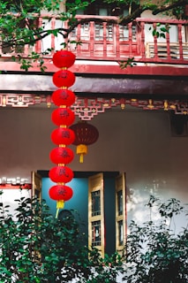 Red lanterns with Chinese characters hang vertically, surrounded by lush green plants and traditional architectural elements. A wooden window with intricate designs is partially open.