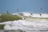 A flock of small birds flies in unison over turbulent ocean waves. The sky is a clear blue, providing a striking contrast to the deep green sea. The birds are captured mid-flight, with their wings spread wide and bodies angled forward, as they navigate the breezy coastal environment.