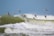 A flock of small birds flies in unison over turbulent ocean waves. The sky is a clear blue, providing a striking contrast to the deep green sea. The birds are captured mid-flight, with their wings spread wide and bodies angled forward, as they navigate the breezy coastal environment.