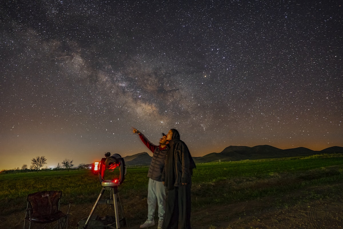 A person beside a tripod-mounted telescope in a field at night pointing up at the Milky Way, a small red light beside them