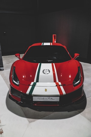 A high-performance red sports car is parked indoors, featuring a sleek design with distinctive red, white, and green racing stripes on the hood. The car has a prominent emblem on the front and a license plate that reads 'Scuderia Praha'. The vehicle is illuminated under studio lighting, emphasizing its aerodynamic shape and glossy finish.