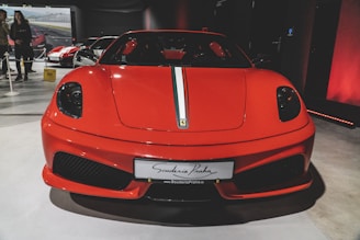 A sleek, red sports car is prominently displayed indoors with a white and green stripe running down its center. The car's front features distinctive headlights and a low aerodynamic profile. In the background, there are other cars and a couple of people observing them.