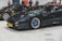 A sleek, dark-colored sports car with a yellow license plate labeled 'LEGENDY' is parked indoors, with several people visible in the background. Other exotic cars are parked nearby, including a black car with white text and details.
