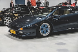 A sleek, dark-colored sports car with a yellow license plate labeled 'LEGENDY' is parked indoors, with several people visible in the background. Other exotic cars are parked nearby, including a black car with white text and details.