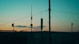 Several antennas and communication equipment are silhouetted against a gradient sky. The sky transitions from blue at the top to a warm orange and yellow near the horizon, suggesting either sunrise or sunset. The scene conveys an industrial setting with a focus on telecommunications.