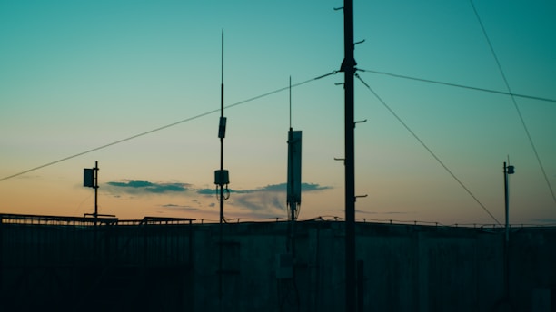 Several antennas and communication equipment are silhouetted against a gradient sky. The sky transitions from blue at the top to a warm orange and yellow near the horizon, suggesting either sunrise or sunset. The scene conveys an industrial setting with a focus on telecommunications.