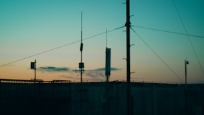 Several antennas and communication equipment are silhouetted against a gradient sky. The sky transitions from blue at the top to a warm orange and yellow near the horizon, suggesting either sunrise or sunset. The scene conveys an industrial setting with a focus on telecommunications.