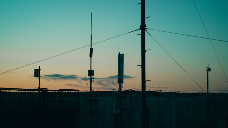 Several antennas and communication equipment are silhouetted against a gradient sky. The sky transitions from blue at the top to a warm orange and yellow near the horizon, suggesting either sunrise or sunset. The scene conveys an industrial setting with a focus on telecommunications.
