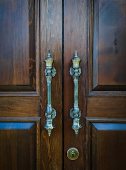 A pair of ornate metallic door handles is mounted on a polished wooden door. The handles feature intricate designs and are positioned symmetrically. Below the handles, a keyhole is visible.