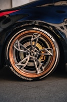 A close-up of a car's wheel featuring a high-performance tire with the brand name 'Hankook' visible. The wheel has a sleek, modern design with a metallic finish and copper-colored rim. The brake caliper is visible inside, adding to the sporty aesthetic. The car's body is dark and glossy, reflecting the surroundings.