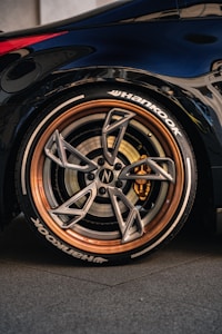 A close-up of a car's wheel featuring a high-performance tire with the brand name 'Hankook' visible. The wheel has a sleek, modern design with a metallic finish and copper-colored rim. The brake caliper is visible inside, adding to the sporty aesthetic. The car's body is dark and glossy, reflecting the surroundings.